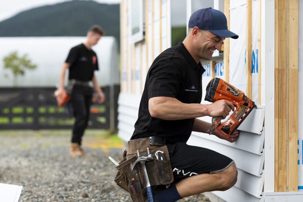 experienced builder using a nail gun to secure weatherboards to an exterior wall, while an apprentice observes and learns.
