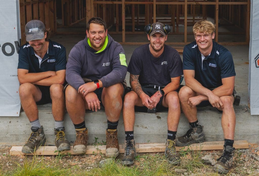 four building apprentices sitting on the concrete slab of a house under construction, smiling as they train to become qualified builders.